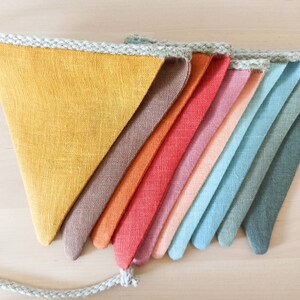 May include: A bunting banner featuring a series of triangular fabric flags in a gradient of colors including mustard yellow, brown, orange, red, pink, peach, light blue, teal, and dark green, all attached to a thick, braided light gray rope.