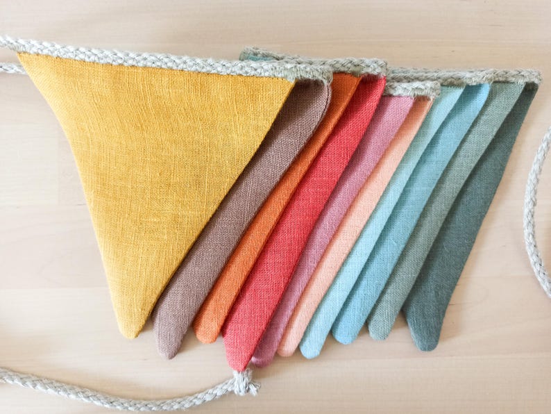 May include: A bunting banner featuring a series of triangular fabric flags in a gradient of colors including mustard yellow, brown, orange, red, pink, peach, light blue, teal, and dark green, all attached to a thick, braided light gray rope.