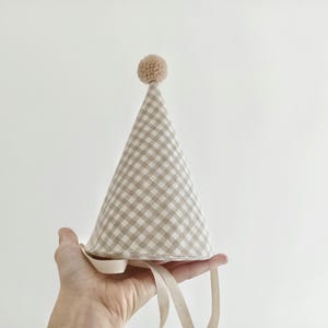 Può includere: Un cappello da festa conico con un motivo a quadretti vichy beige e bianco. Il cappello è sormontato da un piccolo pompon rotondo beige e include due lunghi nastri di tessuto piatti e chiari attaccati alla base per fissare l'oggetto.