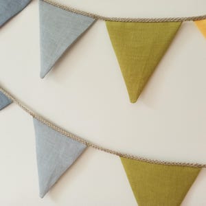 May include: A decorative bunting banner featuring triangular fabric flags in shades of light blue, olive green, and yellow. The flags are attached to a thick, natural-colored twisted jute rope, creating two distinct rows of hanging pennants.