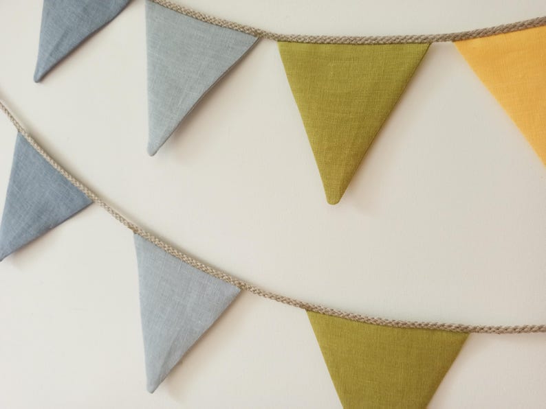 May include: A decorative bunting banner featuring triangular fabric flags in shades of light blue, olive green, and yellow. The flags are attached to a thick, natural-colored twisted jute rope, creating two distinct rows of hanging pennants.