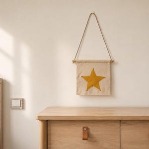 Peut inclure: Une décoration murale beige avec une grande étoile jaune. La bannière est suspendue par une cheville en bois et une corde naturelle. L'image montre également une commode en bois avec des poignées en cuir et un interrupteur.