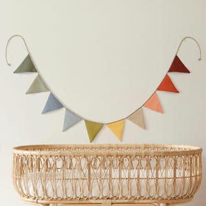 May include: A decorative bunting banner featuring ten triangular fabric flags in a gradient of colors including olive green, sage, blue, grey, mustard yellow, beige, peach, orange, and dark red, suspended on a thin cord in a curved shape.