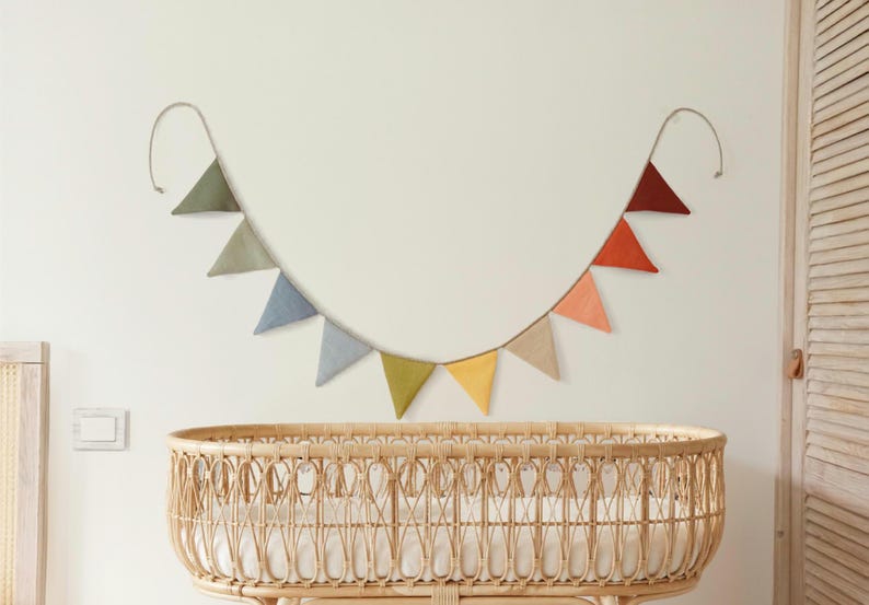 May include: A decorative bunting banner featuring ten triangular fabric flags in a gradient of colors including olive green, sage, blue, grey, mustard yellow, beige, peach, orange, and dark red, suspended on a thin cord in a curved shape.