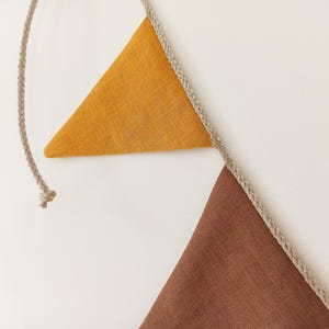 May include: A decorative bunting banner featuring two triangular fabric flags attached to a braided beige rope. One flag is mustard yellow and the other is a deep brown. The fabric has a visible woven texture, and the rope extends beyond the flags.
