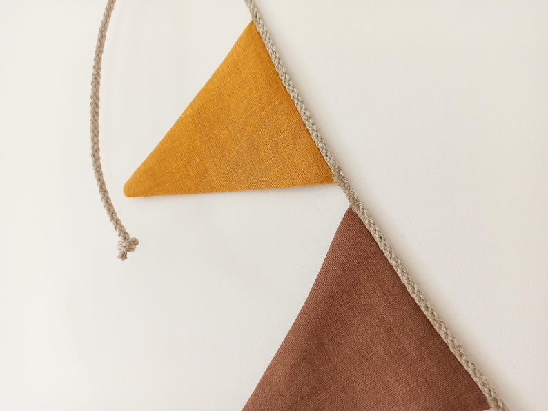 May include: A decorative bunting banner featuring two triangular fabric flags attached to a braided beige rope. One flag is mustard yellow and the other is a deep brown. The fabric has a visible woven texture, and the rope extends beyond the flags.
