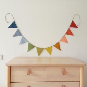 May include: A decorative bunting banner featuring a series of triangular fabric flags in a gradient of colors, including shades of blue, green, yellow, orange, and red, all suspended from a single cord that hangs in a curved, U-shaped line.