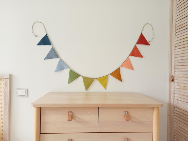 May include: A decorative bunting banner featuring a series of triangular fabric flags in a gradient of colors, including shades of blue, green, yellow, orange, and red, all suspended from a single cord that hangs in a curved, U-shaped line.