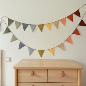 May include: A double-layered fabric bunting banner featuring triangular flags in muted tones of olive green, slate blue, mustard yellow, beige, terracotta, and deep maroon, all suspended from a thin cord arranged in two sweeping, overlapping arcs.