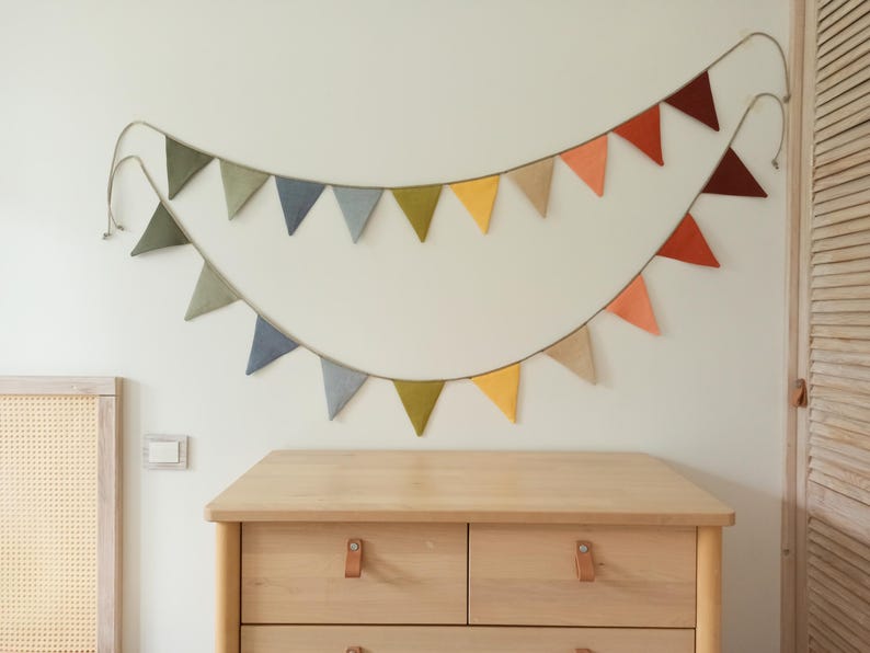 May include: A double-layered fabric bunting banner featuring triangular flags in muted tones of olive green, slate blue, mustard yellow, beige, terracotta, and deep maroon, all suspended from a thin cord arranged in two sweeping, overlapping arcs.