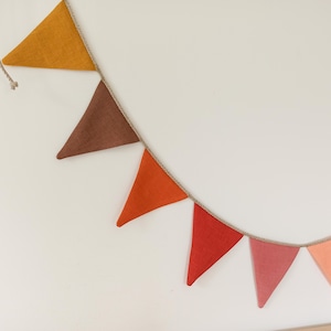 May include: A decorative bunting banner featuring a series of triangular fabric flags in shades of mustard yellow, brown, orange, red, dusty pink, peach, and light blue, all attached to a thin, natural-colored twisted rope string for hanging.