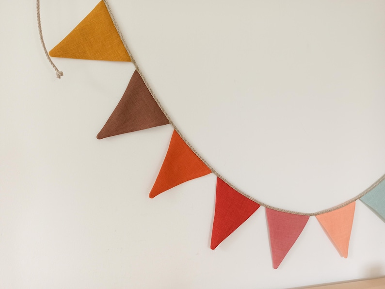 May include: A decorative bunting banner featuring a series of triangular fabric flags in shades of mustard yellow, brown, orange, red, dusty pink, peach, and light blue, all attached to a thin, natural-colored twisted rope string for hanging.