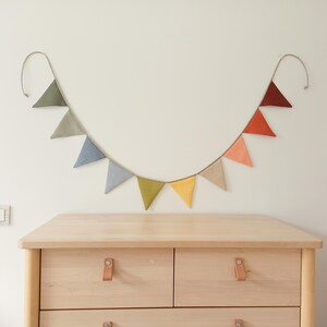 May include: A fabric bunting banner featuring ten triangular flags in a gradient of colors including olive green, sage, slate blue, light blue, lime green, yellow, beige, peach, orange, and dark red, suspended on a thin cord against a wall.