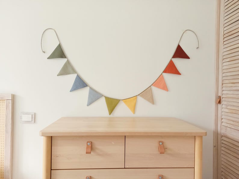 May include: A fabric bunting banner featuring ten triangular flags in a gradient of colors including olive green, sage, slate blue, light blue, lime green, yellow, beige, peach, orange, and dark red, suspended on a thin cord against a wall.