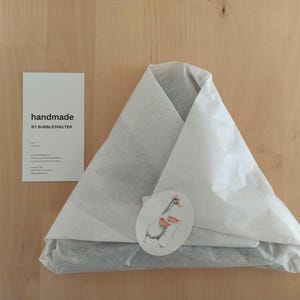 May include: A triangular package wrapped in white tissue paper, sealed with a round sticker featuring a goose illustration. Beside it is a white rectangular card with the black text "handmade BY BUBBLESHELTER" and additional contact information.