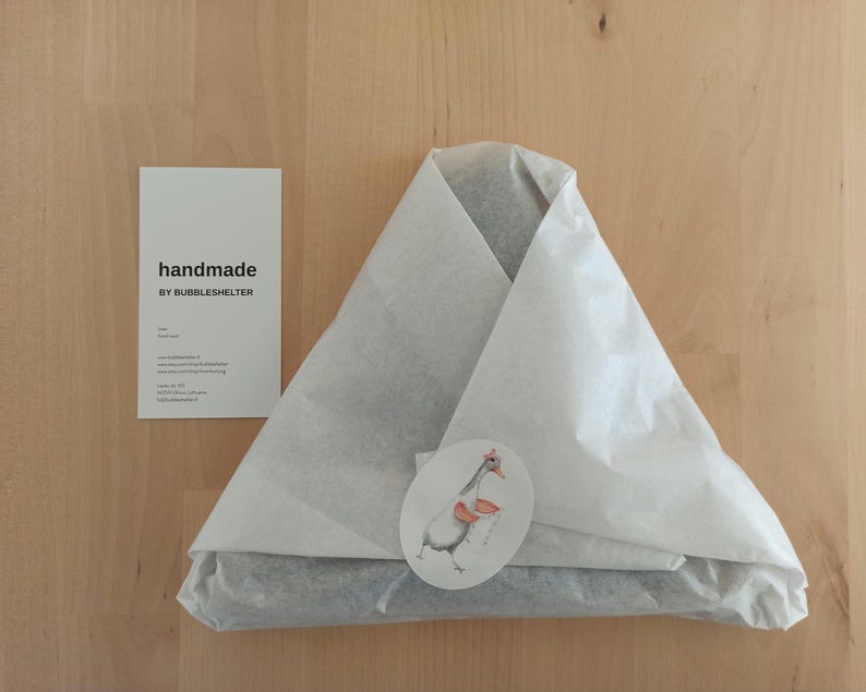 May include: A triangular package wrapped in white tissue paper, sealed with a round sticker featuring a goose illustration. Beside it is a white rectangular card with the black text "handmade BY BUBBLESHELTER" and additional contact information.