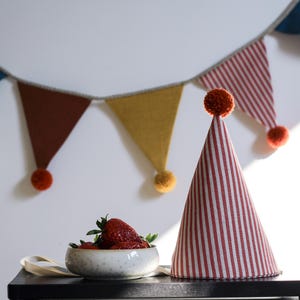 Può includere: Una scena festosa con un cappello da festa a righe rosse e bianche, una ciotola di fragole e una ghirlanda colorata. La ghirlanda ha bandiere triangolari blu, marroni e gialle, con pompon.