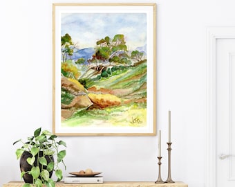 Può includere: Un dipinto ad acquerello di un paesaggio con colline verdi, alberi e un cielo blu. Il dipinto è incorniciato in una cornice di legno chiaro e appeso a una parete bianca.
