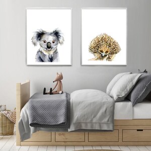 May include: Two watercolor animal prints, one featuring a koala and the other a spiny echidna. The prints are framed in white and displayed on a white wall above a wooden bed with gray bedding.