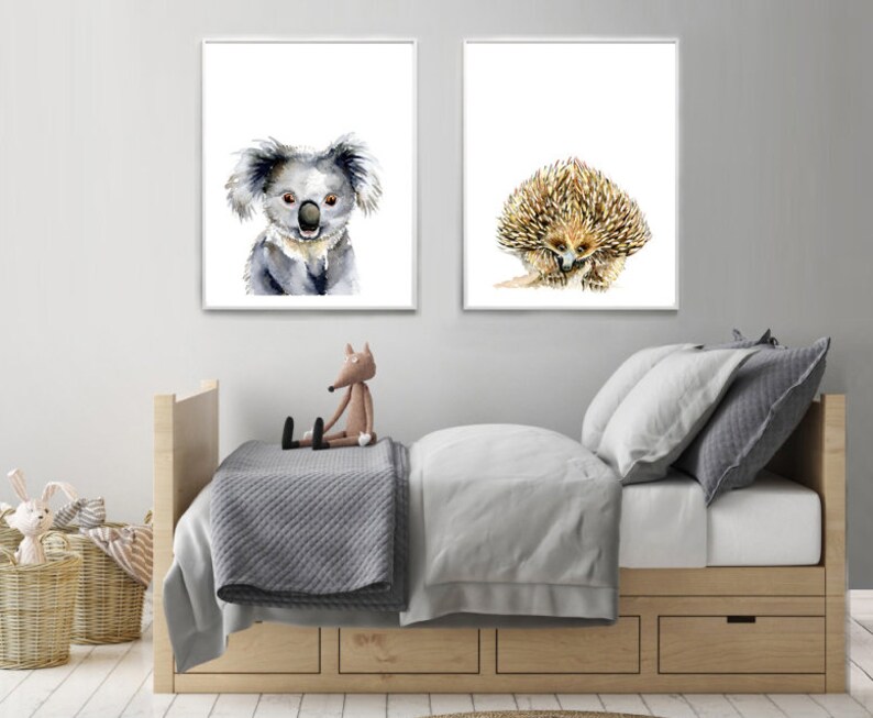 May include: Two watercolor animal prints, one featuring a koala and the other a spiny echidna. The prints are framed in white and displayed on a white wall above a wooden bed with gray bedding.