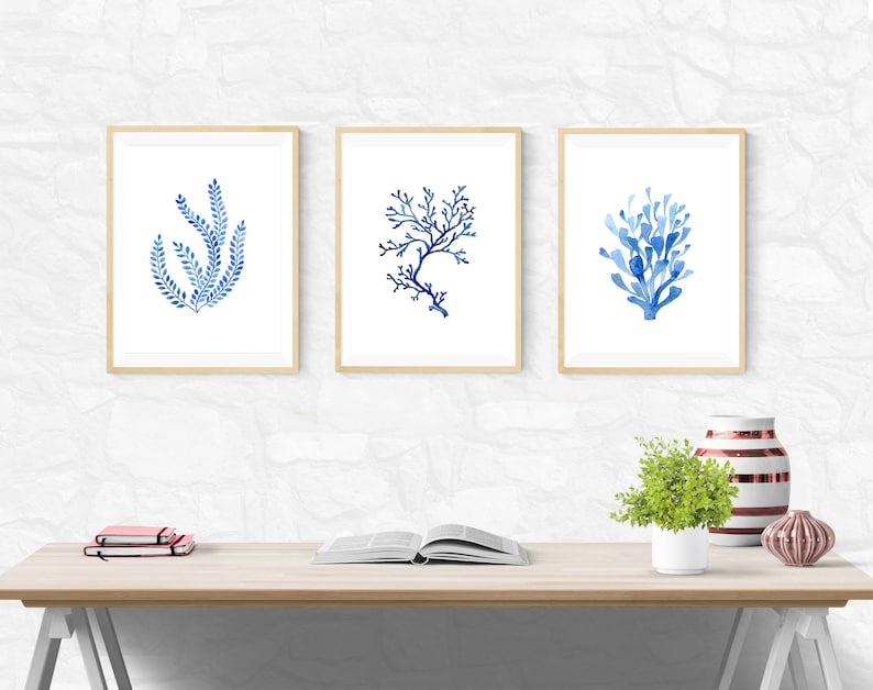 Puede incluir: Tres impresiones enmarcadas en acuarela con dise&ntilde;os de coral y algas marinas azules. La obra de arte se exhibe sobre un escritorio de madera clara con un fondo blanco. Una peque&ntilde;a planta, libros y jarrones decorativos tambi&eacute;n est&aacute;n en el escritorio.