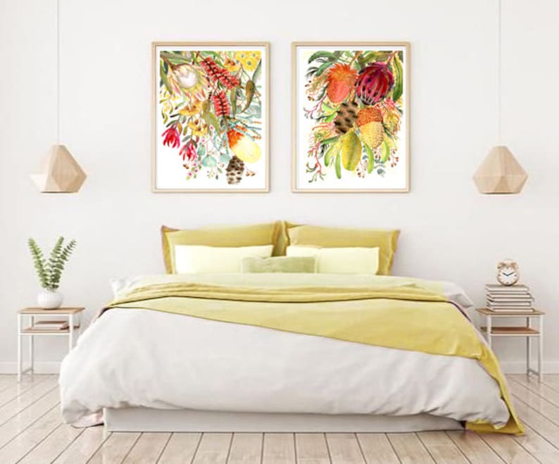 Puede incluir: Dos acuarelas enmarcadas de arreglos florales con flores rojas, amarillas y verdes. Las pinturas est&aacute;n colgadas en una pared blanca sobre una colcha de cama amarilla.