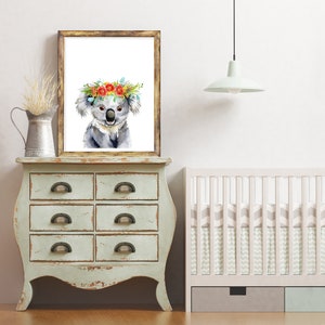 Puede incluir: Una pintura de acuarela de un koala con una corona de flores. El koala es gris y tiene la nariz y el pecho blancos. La corona est&aacute; hecha de flores rojas, naranjas y amarillas. La pintura est&aacute; enmarcada en un marco de madera r&uacute;stico y est&aacute; colgada en una pared blanca.