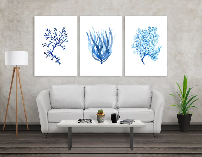 Puede incluir: Tres impresiones enmarcadas en acuarela con dise&ntilde;os de coral y algas marinas azules. La obra de arte se exhibe sobre un sof&aacute; blanco, con una l&aacute;mpara moderna y una planta en maceta en la habitaci&oacute;n. Las impresiones a&ntilde;aden un tema costero a la habitaci&oacute;n.