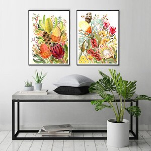 Puede incluir: Dos pinturas de acuarela enmarcadas de coloridas flores nativas australianas, incluyendo tonos rojos, amarillos y verdes, expuestas en un estante de concreto con una planta en maceta y una pila de libros.