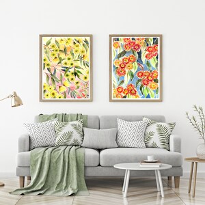 以下が含まれることがあります： 花柄のアレンジメントを描いた額装された水彩画が2点。左側の絵は、ピンクの背景に緑の葉をつけた黄色の花が描かれている。右側の絵は、青い背景に緑の葉をつけた赤い花が描かれている。絵は、灰色のソファの上に置かれた緑色のクッションのある白い壁に飾られている。