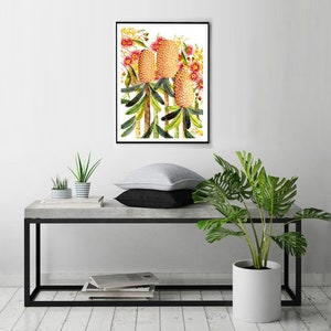 Puede incluir: Una pintura de acuarela enmarcada de tres flores de Banksia amarillas y rosadas con hojas verdes sobre un fondo blanco.