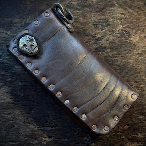 Könnte beinhalten: Eine dunkelbraune Ledergeldbörse mit einer Reihe von Metallnieten entlang der Kante, einem strukturierten silberfarbenen Totenkopf-Emblem in der oberen Ecke und einem schweren Metallring, der am oberen Ende des rechteckigen Accessoires befestigt ist.
