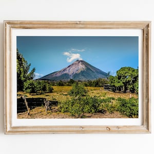 May include: A framed landscape photograph featuring a conical volcano with a small plume of smoke at the peak, set against a clear blue sky. The foreground shows a grassy field with scattered green trees and shrubs under bright daylight.