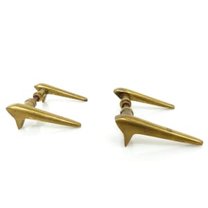 Puede incluir: Dos manijas de puerta de latón con un diseño elegante y moderno. Cada manija presenta una palanca larga y cónica y un extremo puntiagudo distintivo. Las manijas se colocan sobre un fondo blanco, resaltando su acabado dorado pulido.