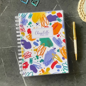 May include: A spiral-bound planner with a white cover featuring a colorful abstract pattern of yellow, orange, red, purple, and green paint strokes and scribbles. The center label reads Chrystelle 2025-2026 in a script font.