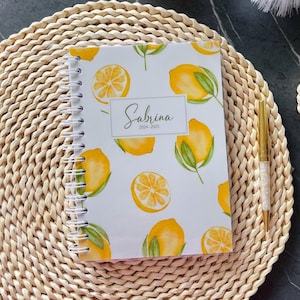 Op de afbeelding: Een spiraalgebonden planner met een witte kaft met een citroenpatroon en de naam "Sabrina" in sierlijke letters. De planner bevat de jaren 2024-2025. Een gouden pen met een kristalachtig ontwerp ligt naast de planner op een geweven placemat.