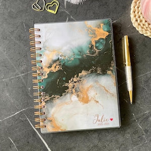 Puede incluir: Un cuaderno con espiral con una cubierta de diseño marmolado verde y dorado. El cuaderno está personalizado con el nombre "Julie" y los años "2023-2026". Un bolígrafo dorado con un cuerpo de cristal descansa junto al cuaderno.