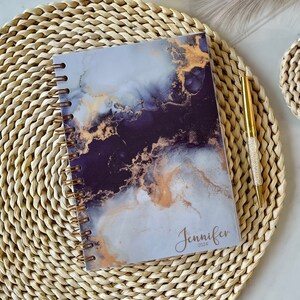 Puede incluir: Un cuaderno de espiral con una cubierta de mármol en tonos grises, negros y dorados. La cubierta tiene el nombre "Jennifer" y el año "2024" impreso.