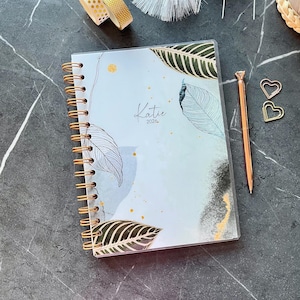 Peut inclure: Un agenda à reliure spirale avec une couverture bleu clair ornée d'illustrations de feuilles vert foncé, de taches dorées et du texte "Katie 2026" en écriture noire. L'agenda est accompagné d'un stylo or rose et deux trombones en forme de cœur.