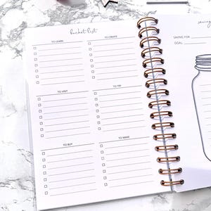 May include: Open notebook with a copper spiral binding. The pages are white with a bucket list layout, including sections for learning, creating, visiting, trying, buying, and making. The words "bucket list" are at the top.