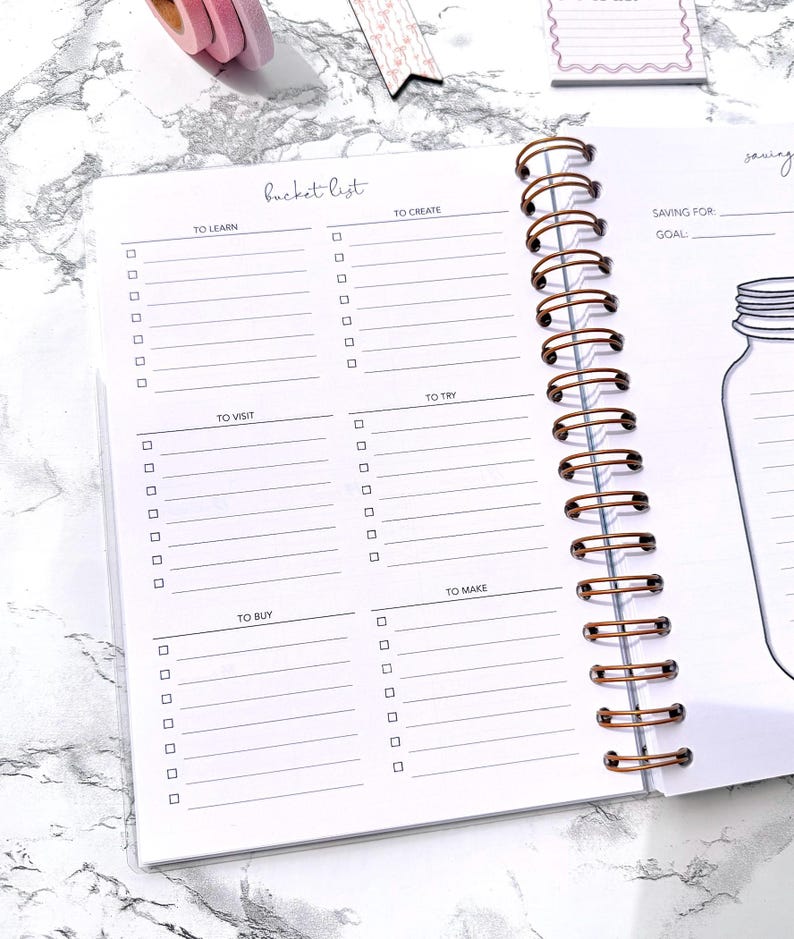 May include: Open notebook with a copper spiral binding. The pages are white with a bucket list layout, including sections for learning, creating, visiting, trying, buying, and making. The words "bucket list" are at the top.