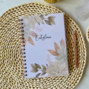 Könnte beinhalten: Ein spiralgebundener Planer mit weißem Einband, der mit Aquarellillustrationen von beigen und salbeigrünen Weihnachtssternen und Blättern verziert ist. Der Text Catalina 2024 ist in schwarzer Schrift gedruckt. Ein roségoldener Stift liegt daneben.