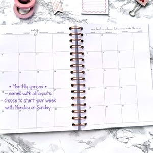 May include: Open spiral-bound planner with a monthly spread layout. The word "may" is handwritten at the top. Text on the page reads "Monthly spread - comes with all layouts - choose to start your week with Monday or Sunday." A rose gold pen and washi tape are visible.