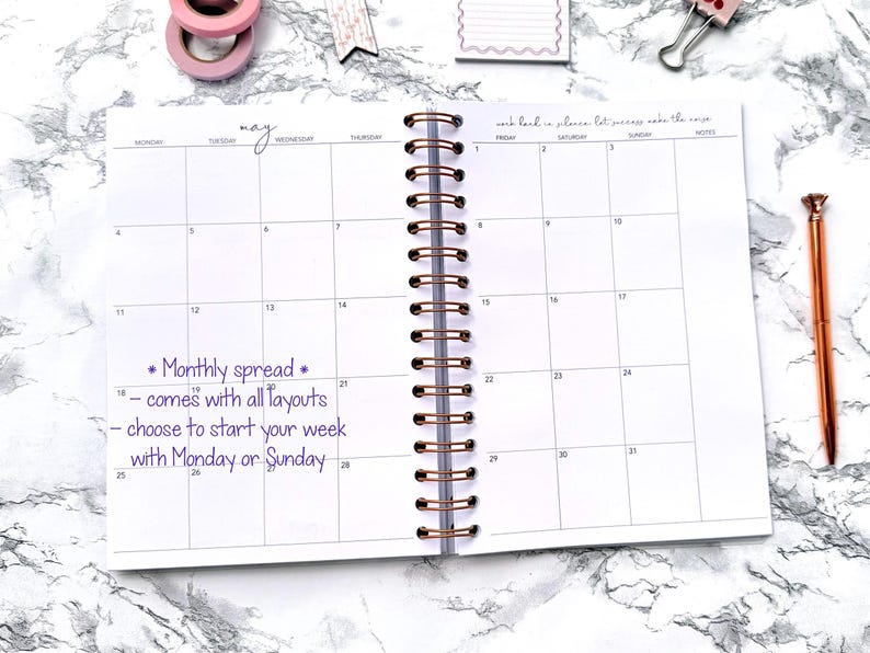 May include: Open spiral-bound planner with a monthly spread layout. The word "may" is handwritten at the top. Text on the page reads "Monthly spread - comes with all layouts - choose to start your week with Monday or Sunday." A rose gold pen and washi tape are visible.
