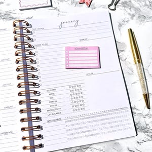 May include: Open spiral-bound planner with a gold pen. The planner is open to a page labeled "January" with sections for goals, tasks, and habits. A pink checklist sticky note is attached.