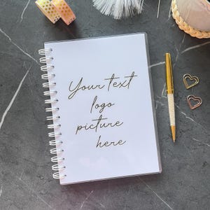 May include: A white spiral-bound notebook with the text "Your text logo picture here" in elegant script. A gold pen with a crystal-like design and two heart-shaped paper clips are next to the notebook. Decorative tape and a small woven item are also visible.