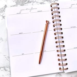 May include: An open planner with a rose gold pen. The planner is white with a spiral binding and the words "year at a glance" at the top. The pen has a diamond-shaped top and is resting on the page.
