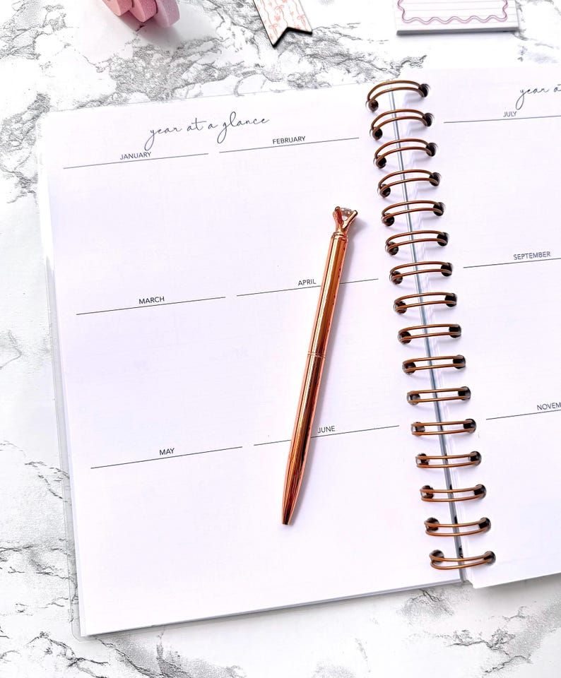 May include: An open planner with a rose gold pen. The planner is white with a spiral binding and the words "year at a glance" at the top. The pen has a diamond-shaped top and is resting on the page.