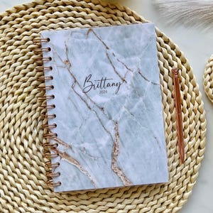 May include: A spiral-bound notebook with a light blue and white marble pattern cover featuring the name Brittany and the year 2024 in black script text, accompanied by a metallic rose gold pen with a diamond-shaped topper on the side.