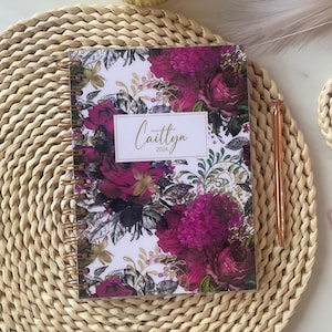 Könnte beinhalten: Ein Spiralplaner mit floralem Design, das tiefrosa und burgunderfarbene Rosen und Hortensien zeigt. Auf dem Cover steht der Name "Caitlyn" und das Jahr "2024" in Gold. Ein goldener Stift liegt neben dem Planer.